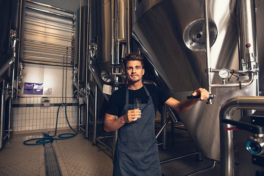 Business Insurance - Young Brewery Owner in Black Shirt and Gray Apron Holds a Beer Sample as He Poses in Front of Large Steel Brewing Tanks