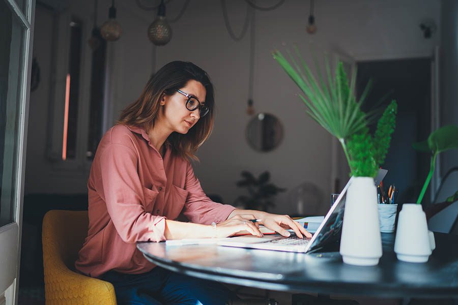 Client Center - Young Woman in Business Casual Clothing and Glasses Uses Her Laptop at a Table by a Window in Her Modern Office Surrounded by Plants