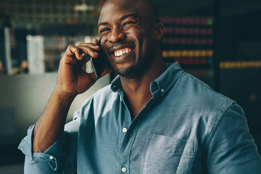 Contact Us - Man Smiling Broadly in a Button-Down Denim Shirt Makes a Phone Call in His Dark Modern Office