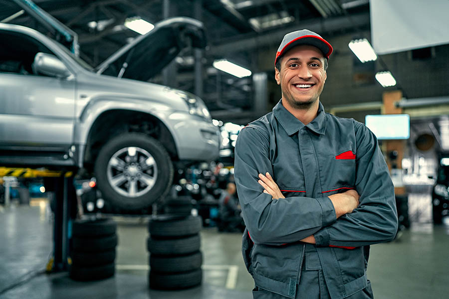 Specialized Business Insurance - Auto Mechanic Smiles and Crosses His Arms With an Elevated Car and Spare Tires Stacked Behind Him in His Large High End Auto Shop